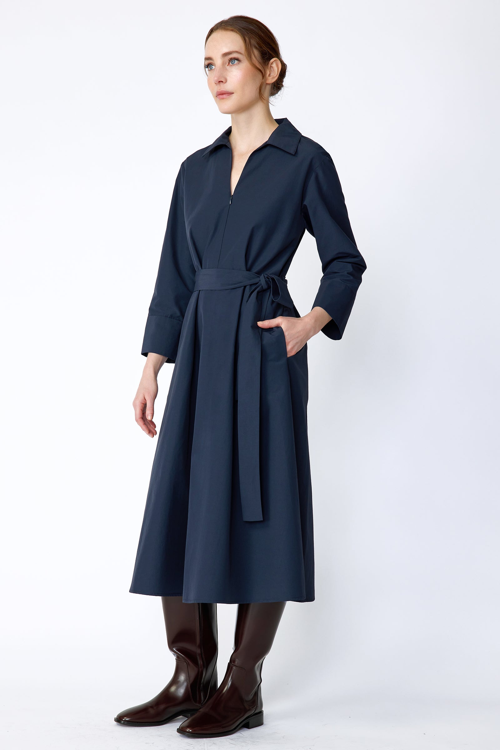 Woman wearing Kal Rieman Sydney Dress in Navy on a white background with brown boots, hand in pocket, arm on the side, side view
