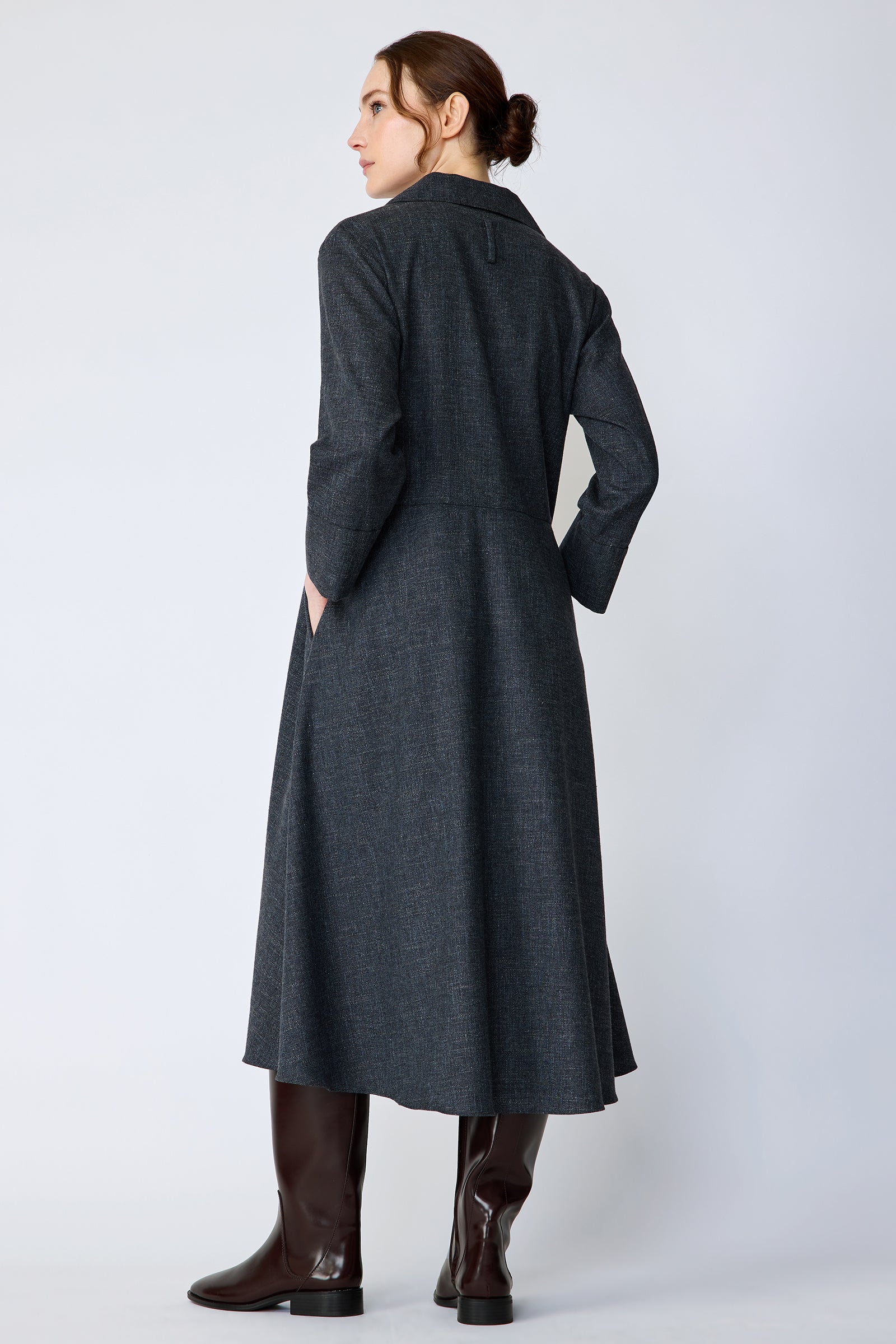 Woman wearing Kal Rieman Sydney Dress in Indigo Denim Wool with brown boots, hands in pockets, looking sideways, full back view