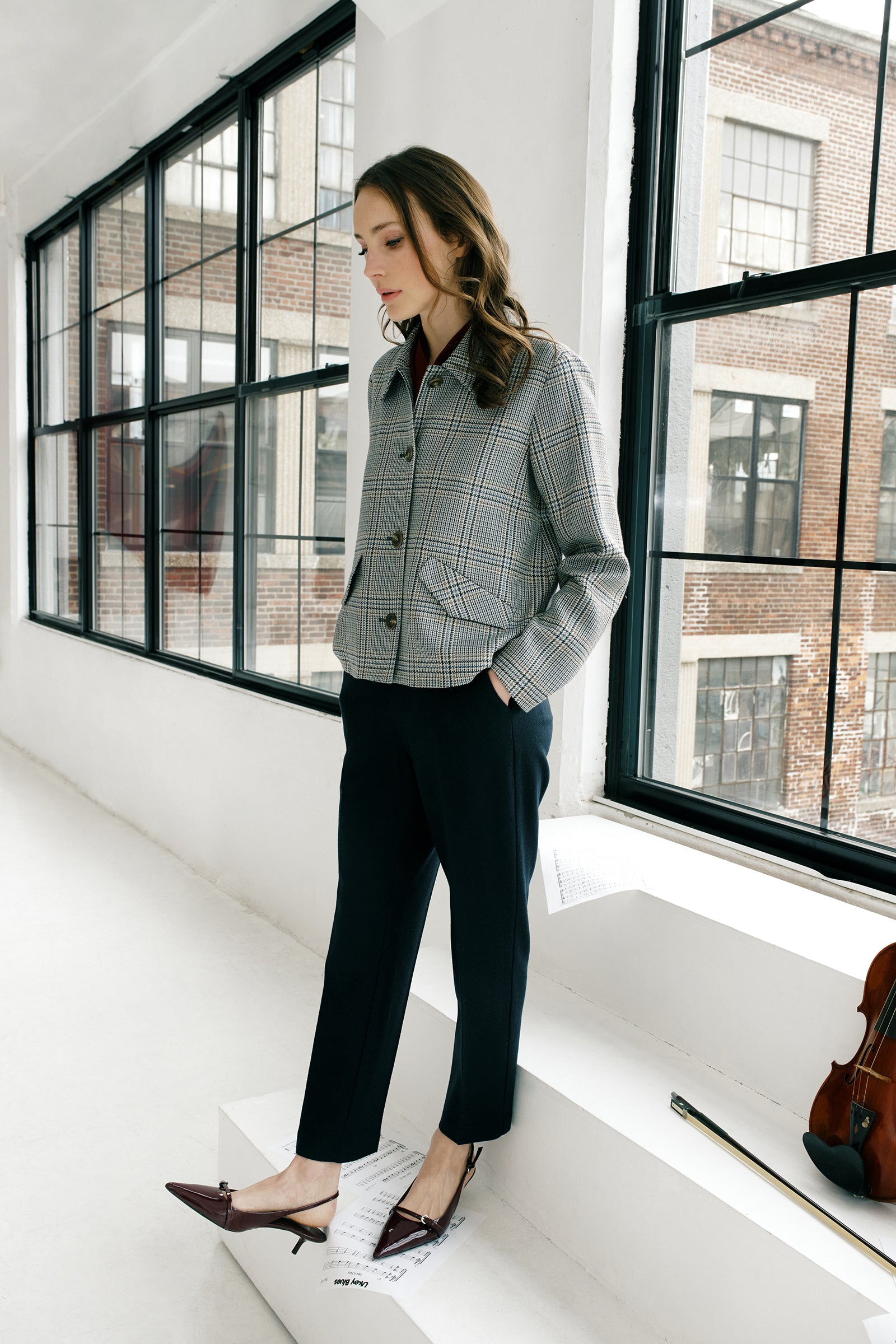 Woman wearing Kal Rieman Sylvie Swing Jacket in Italian Glen Plaid and black pants in a room, descending down steps with a violin in the background arms in pocket look book image