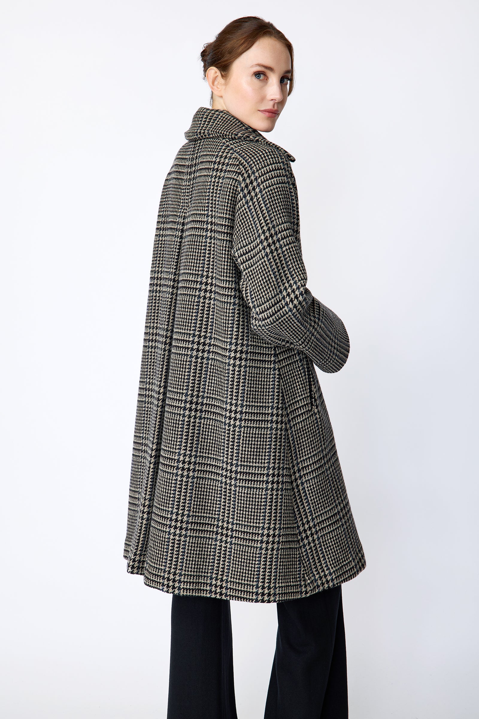 Woman wearing Kal Rieman Venice Coat in Italian Plaid over Kal Rieman Vanessa Bow Blouse in white cotton against a white wall with a red fabric draped over it, hands in pockets, look book image