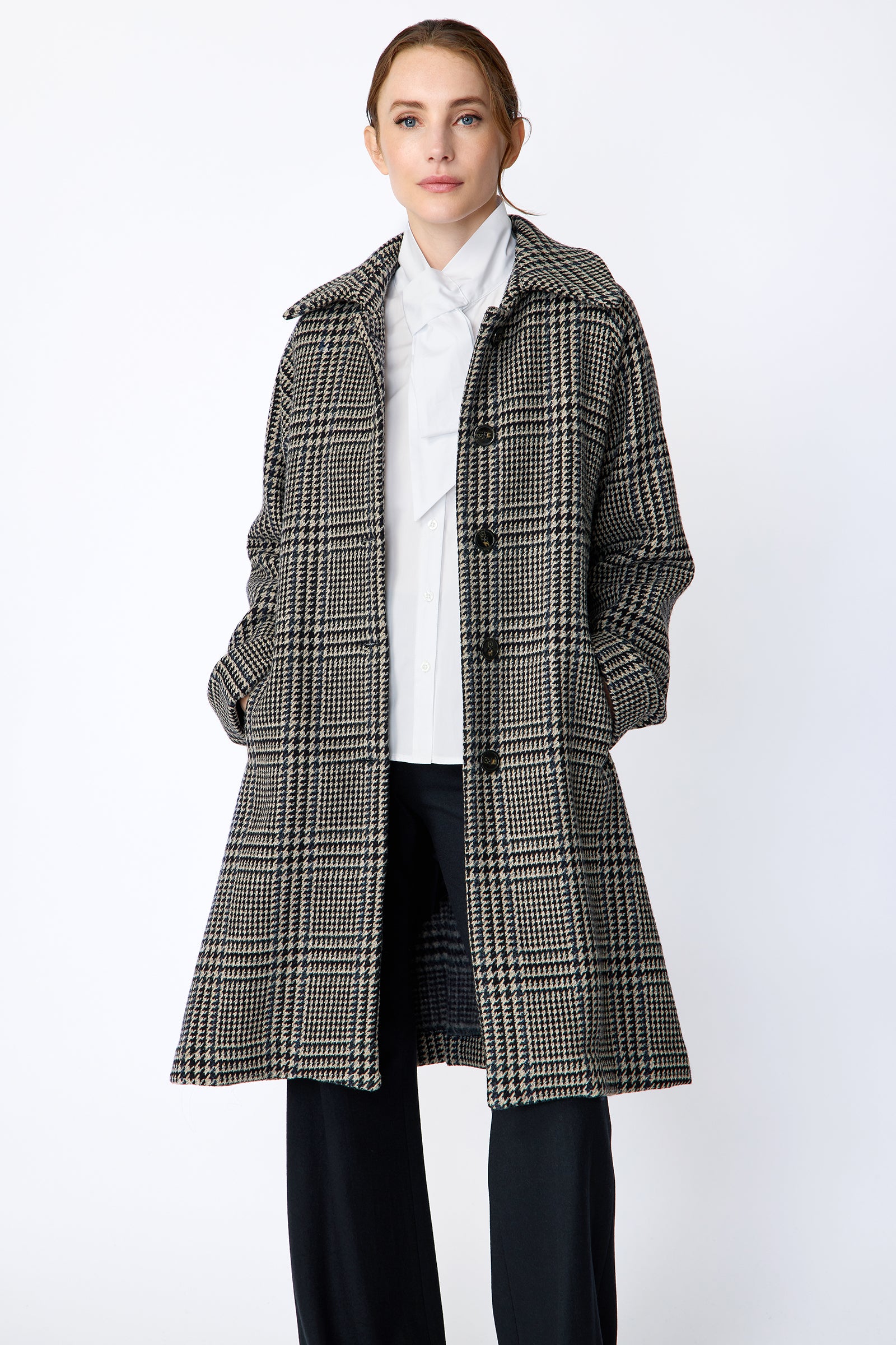 Woman wearing Kal Rieman Venice Coat in Italian Plaid unbuttoned over Kal Rieman Vanessa Bow Blouse in White Cotton with Kal Rieman Felted Jersey Wide Leg pants in Midnight against a white wall, hands in pant pockets
