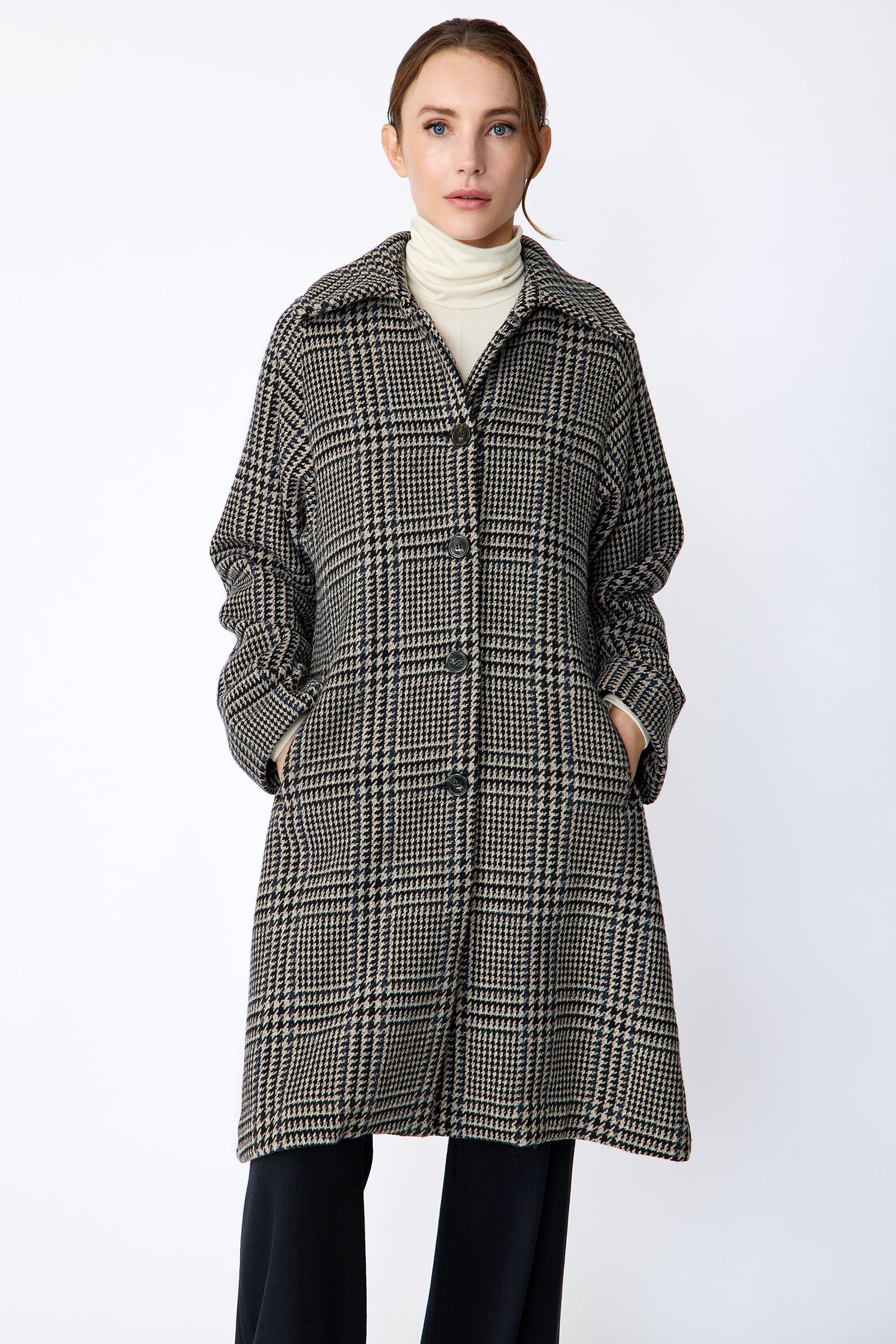 Woman wearing Kal Rieman Venice Coat in Italian Plaid with Kal Rieman Felted Jersey Wide Leg pants in Midnight against a white wall, hands in coat pockets, close view