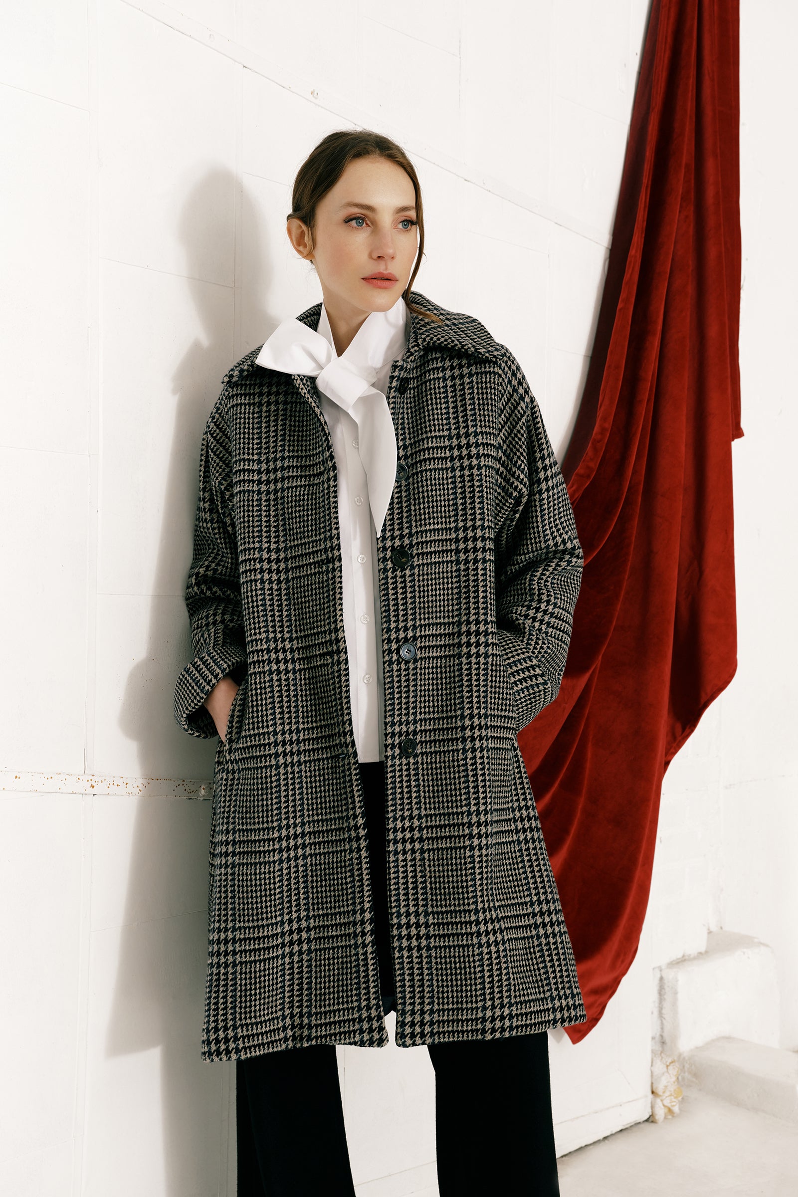 Woman wearing Kal Rieman Venice Coat in Italian Plaid over Kal Rieman Vanessa Bow Blouse in white cotton against a white wall with a red fabric draped over it, hands in pockets, look book image