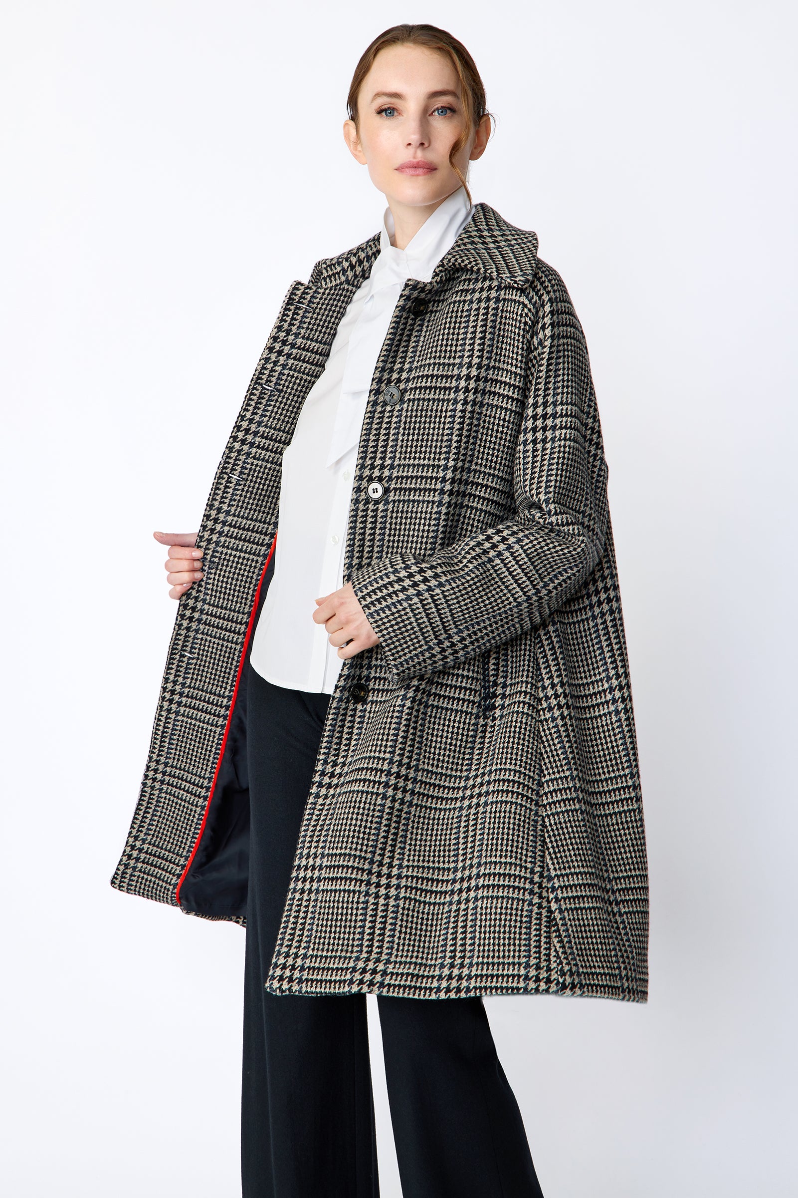 Woman wearing Kal Rieman Venice Coat in Italian Plaid unbuttoned over Kal Rieman Vanessa Bow Blouse in White Cotton with Kal Rieman Felted Jersey Wide Leg pants in Midnight against a white wall, holding coat open