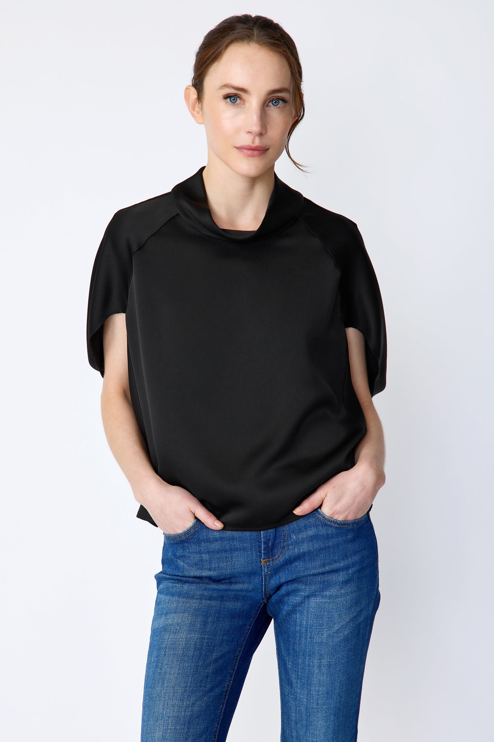 Woman wearing Kal Rieman Angelica Cape Top in Black and blue jeans on a white background, hands in pant pockets