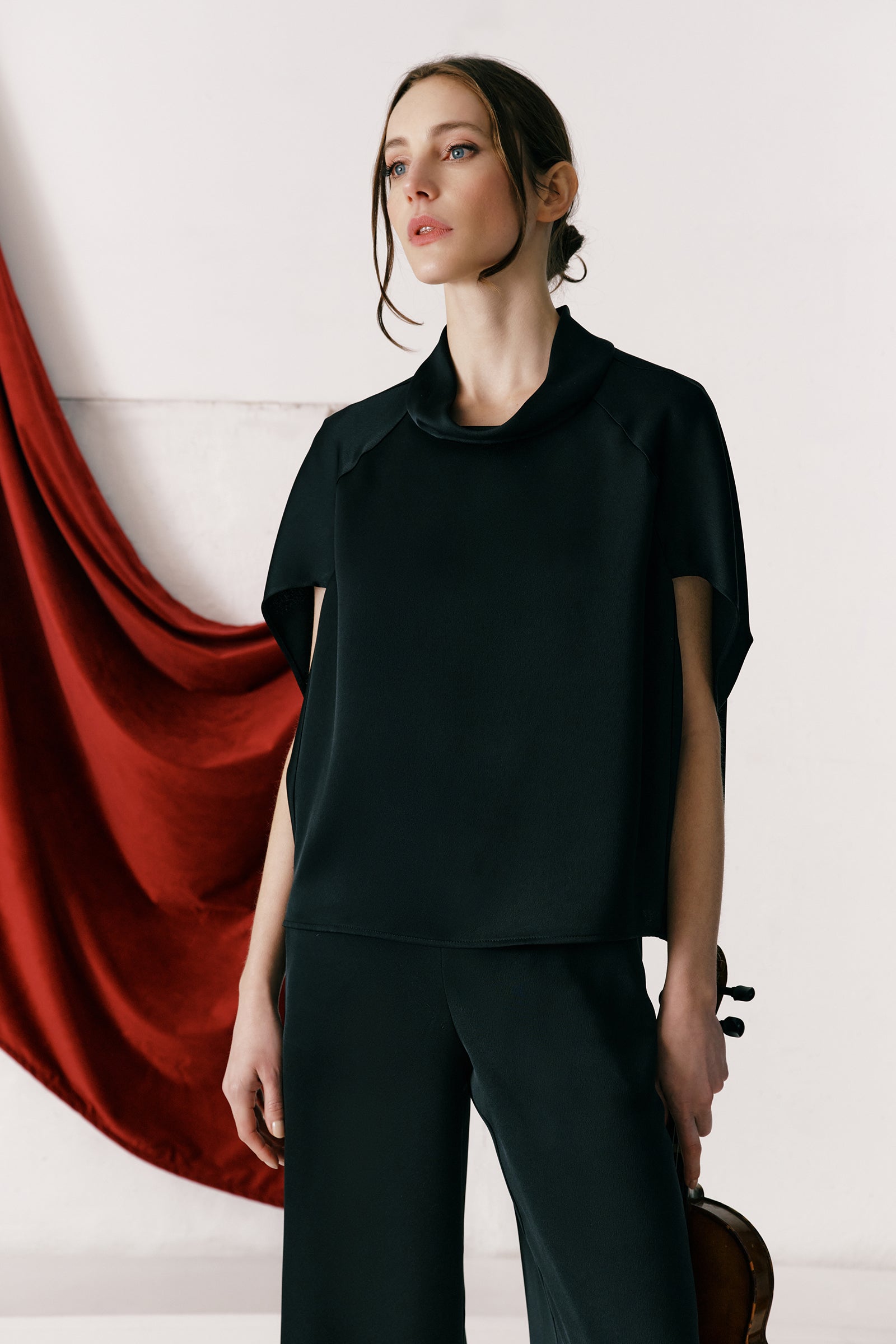 Woman wearing Kal Rieman Angelica Cape Top in Black and blue jeans on a white background, hands in pant pockets