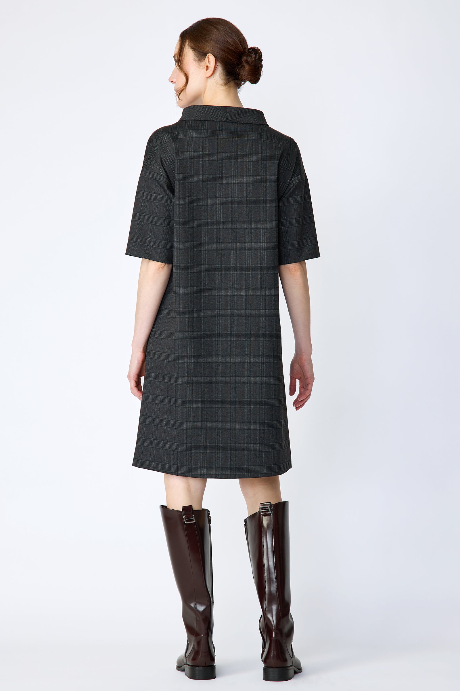 Woman wearing Kal Rieman Avery shift dress in black glen plaid and brown knee-high boots on a white background with arms on the side