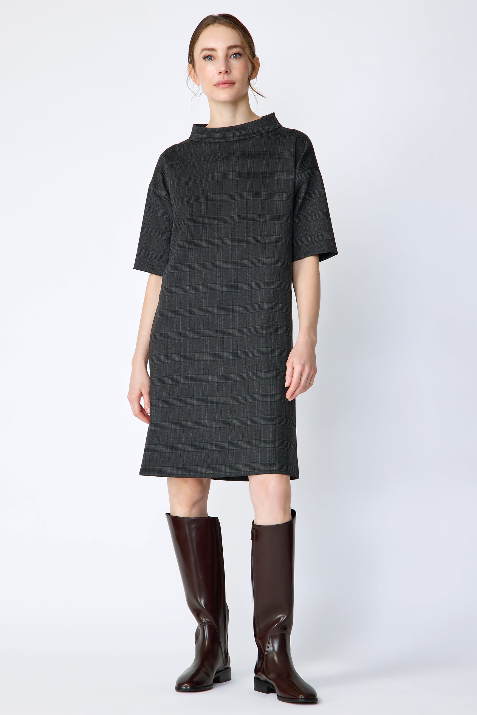 Woman wearing Kal Rieman Avery shift dress in black glen plaid and brown knee-high boots on a white background with arms on the side