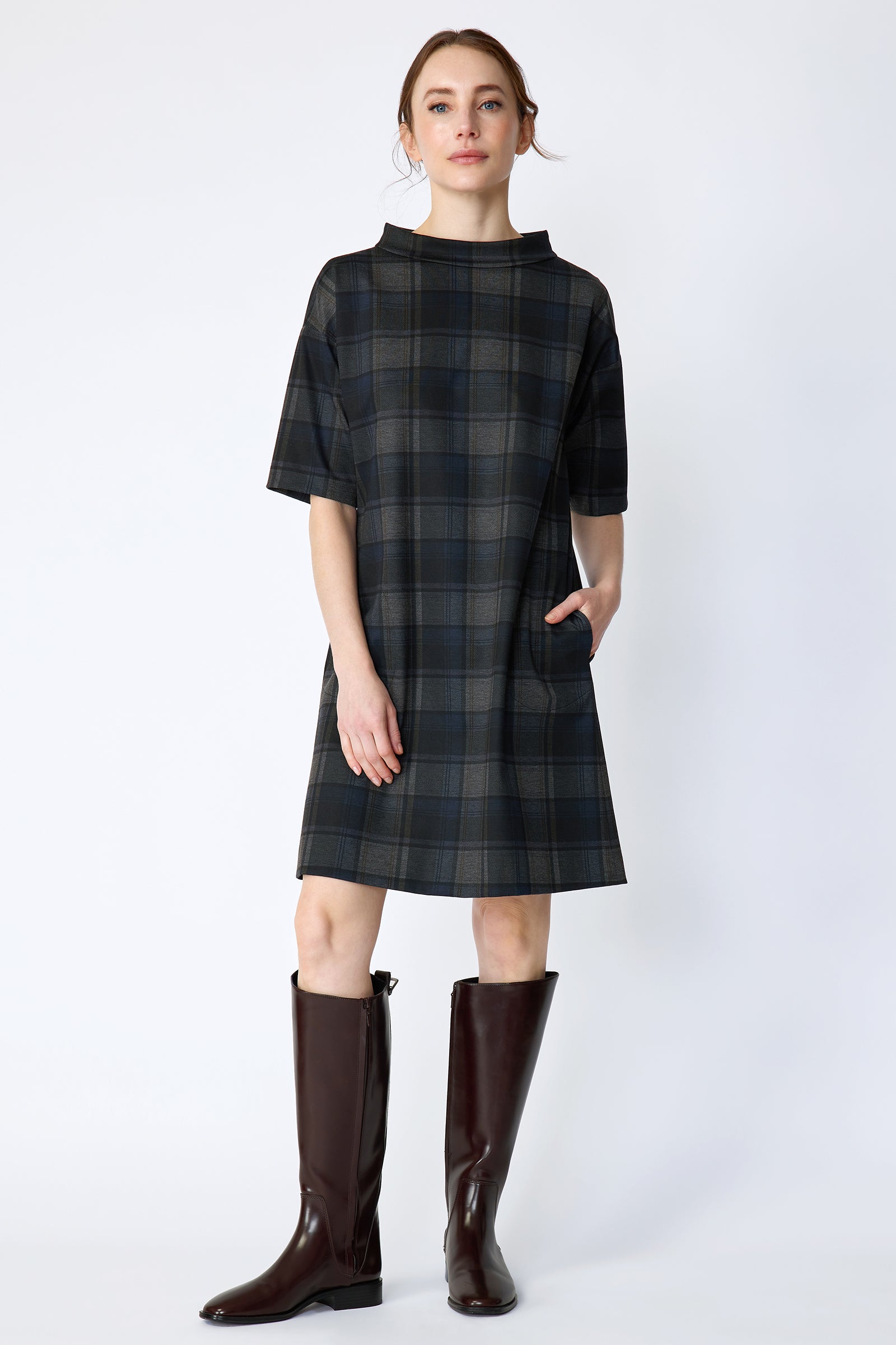Woman wearing Kal Rieman Avery Shift Dress in Novelty Plaid with knee-high boots on a white background with one arm in pocket
