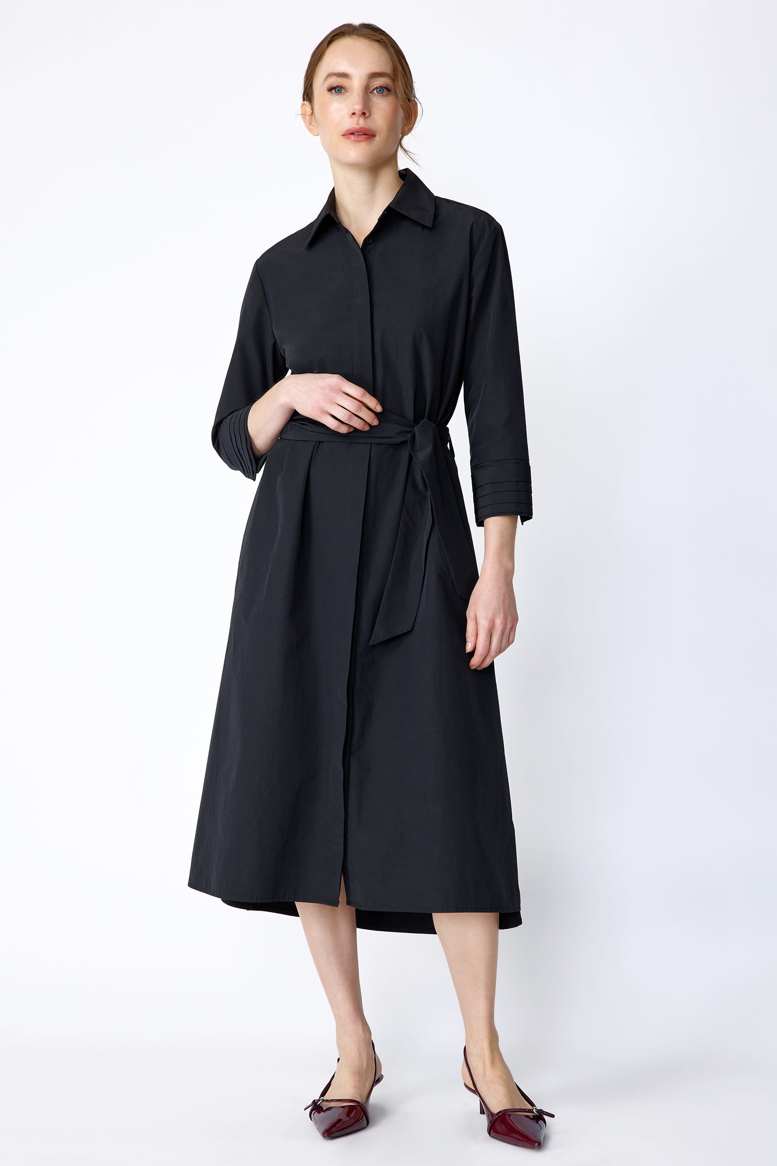 Woman wearing Kal Rieman Carolina Dress in Black Italian Broadcloth with burgundy shoes, against a white background, hand holding belt, arm on the side, full front view