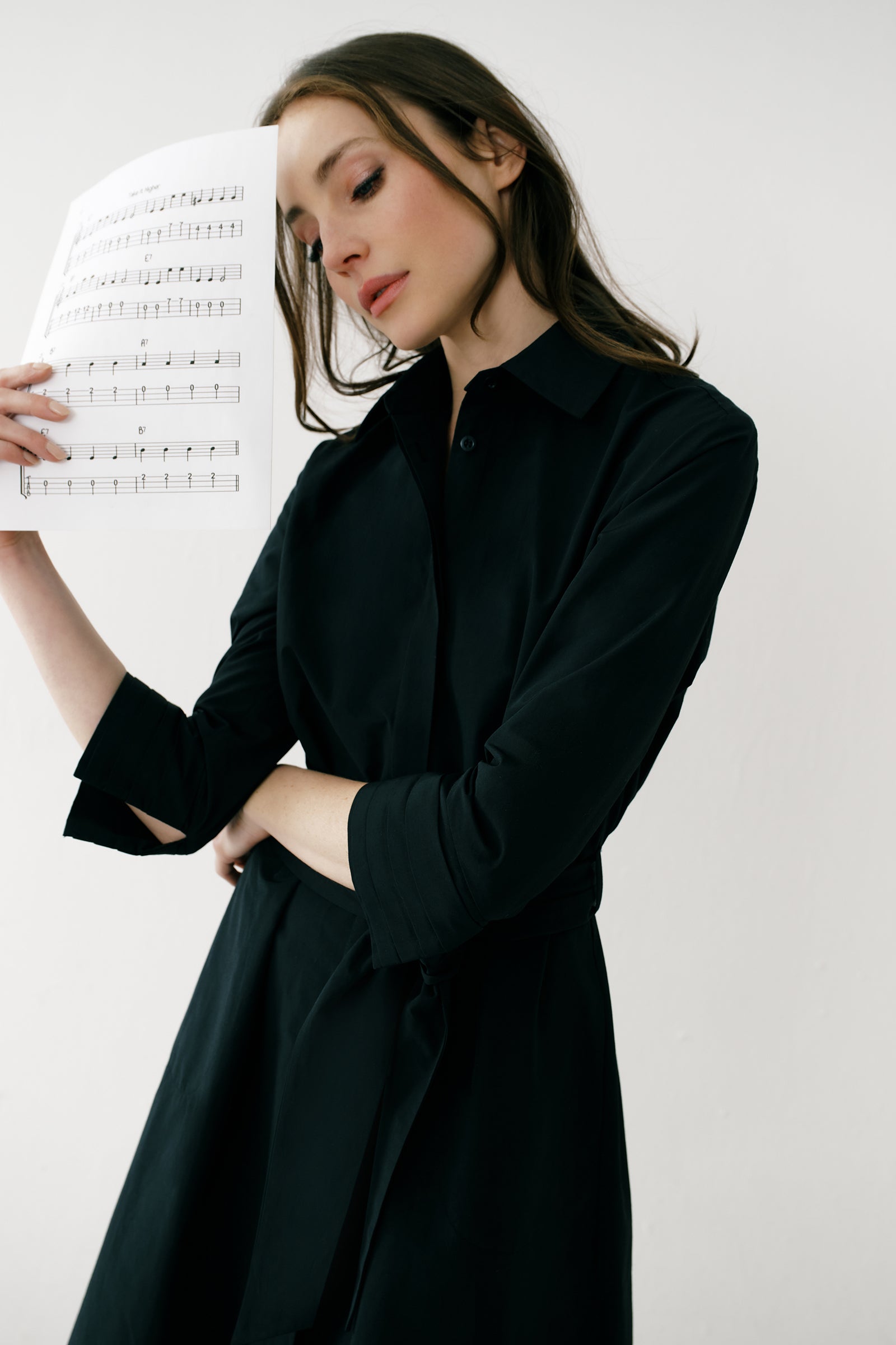 Woman wearing Kal Rieman Carolina Dress in Black Italian Broadcloth with burgundy shoes, against a white background, hand holding belt, arm on the side, full front view