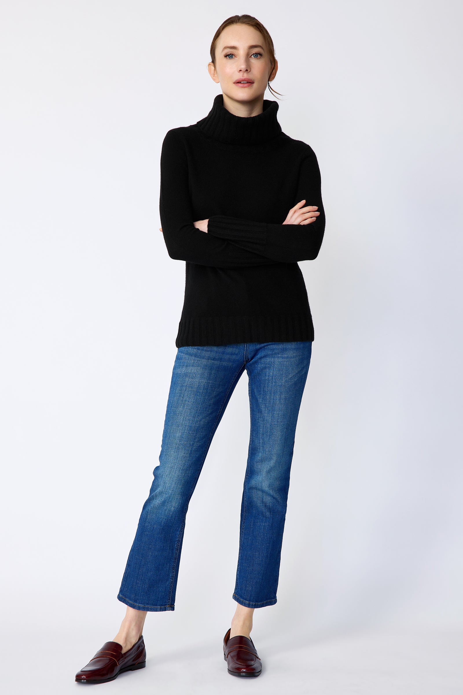Woman wearing a Kal Rieman Cashmere Cown Tneck in black sweater and blue jeans on a white background arms crossed in front