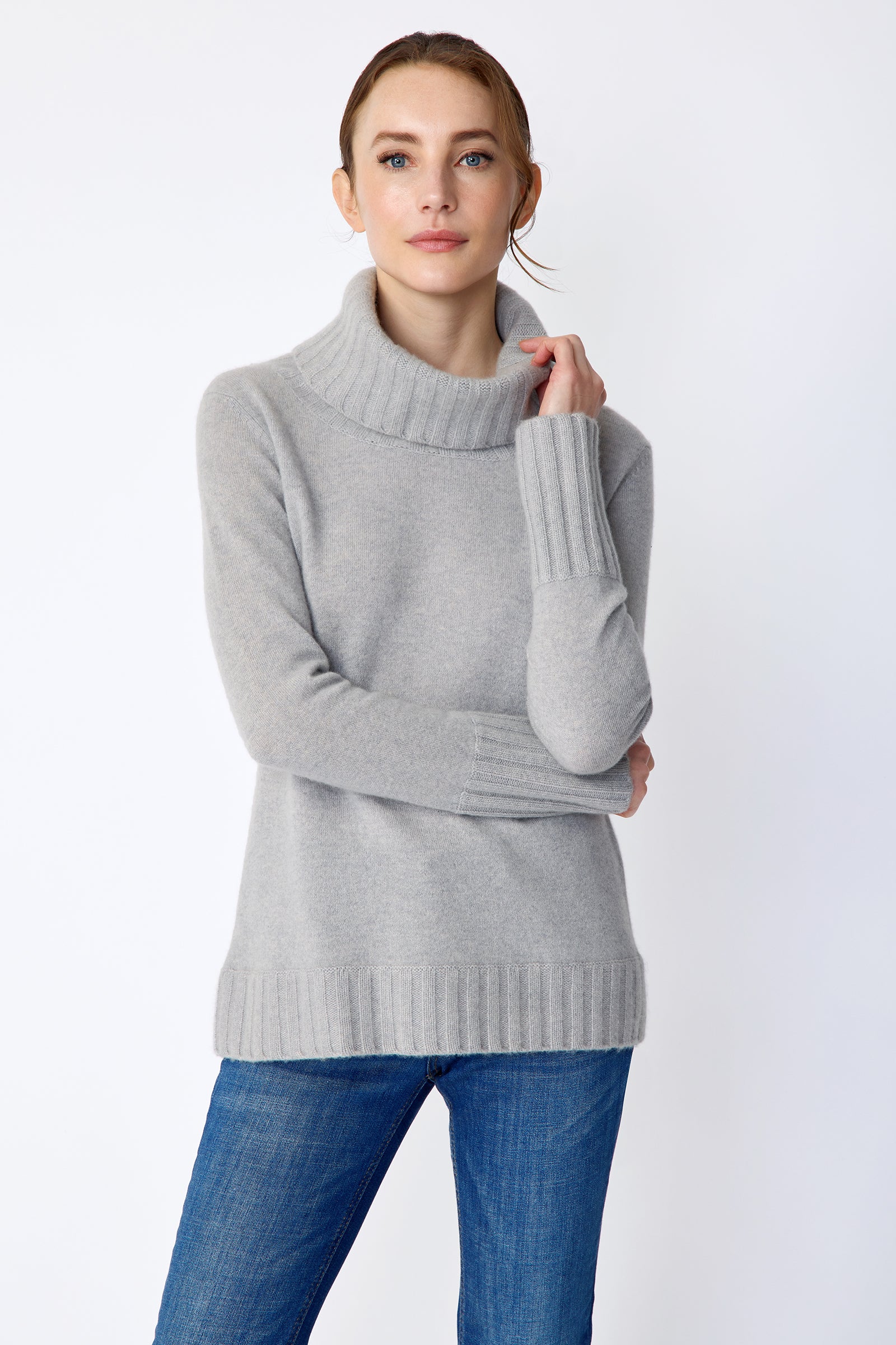 Woman wearing Kal Rieman Cashmere Cowl Tneck in Heather Grey and blue jeans on a white background arms crossed in front, one arm holding collar