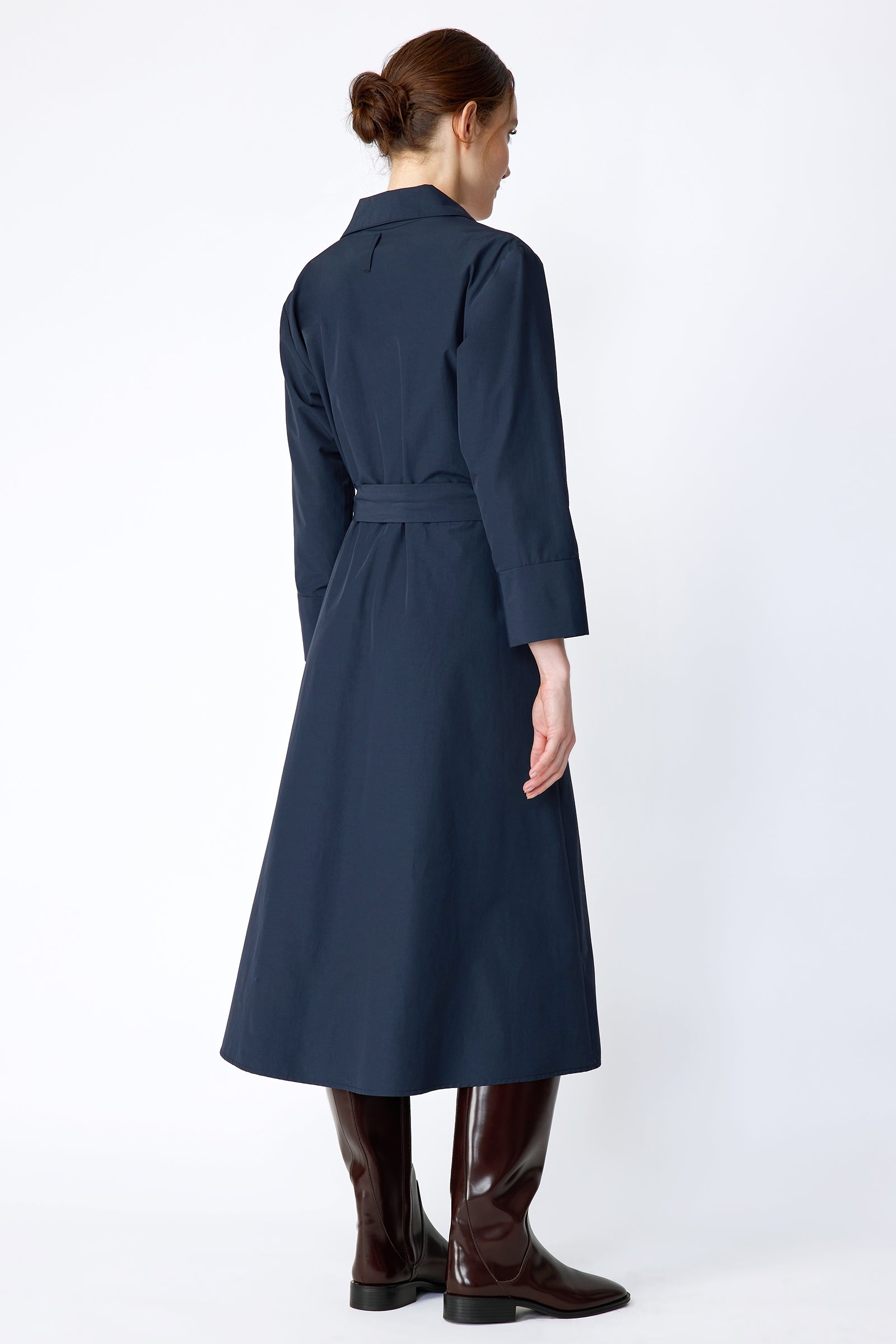 Woman wearing Kal Rieman Sydney Dress in Navy on a white background with brown boots, hands in pockets, in movement, looking down