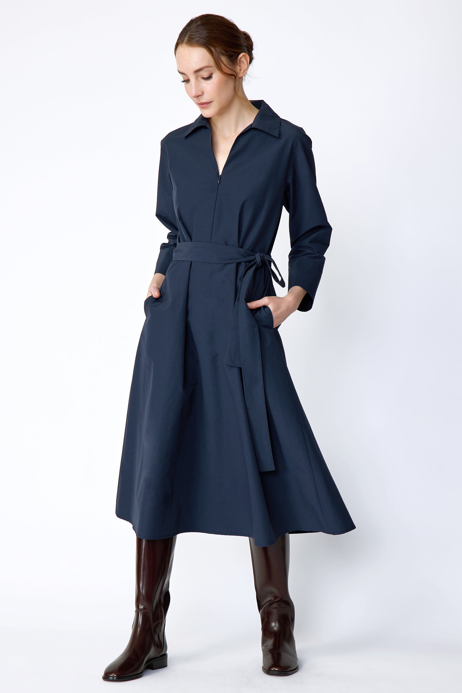 Woman wearing Kal Rieman Sydney Dress in Navy on a white background with brown boots, hands in pockets, in movement, looking down